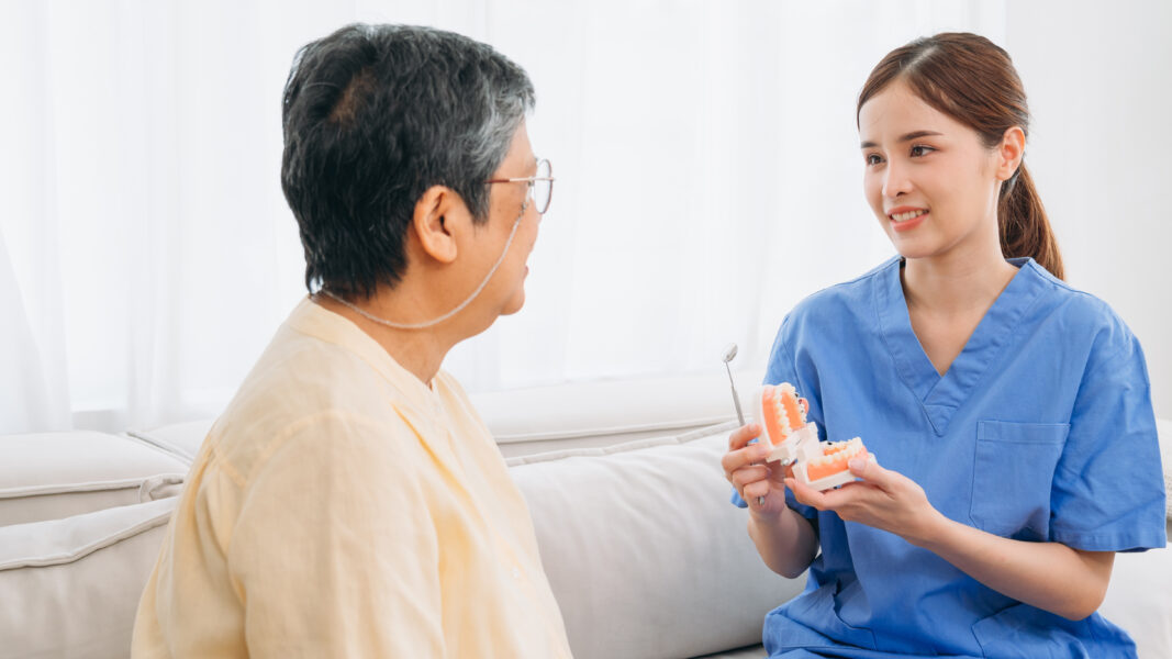 A woman in a blue uniform is holding a toothbrush and talking to an older man. The man is sitting on a couch